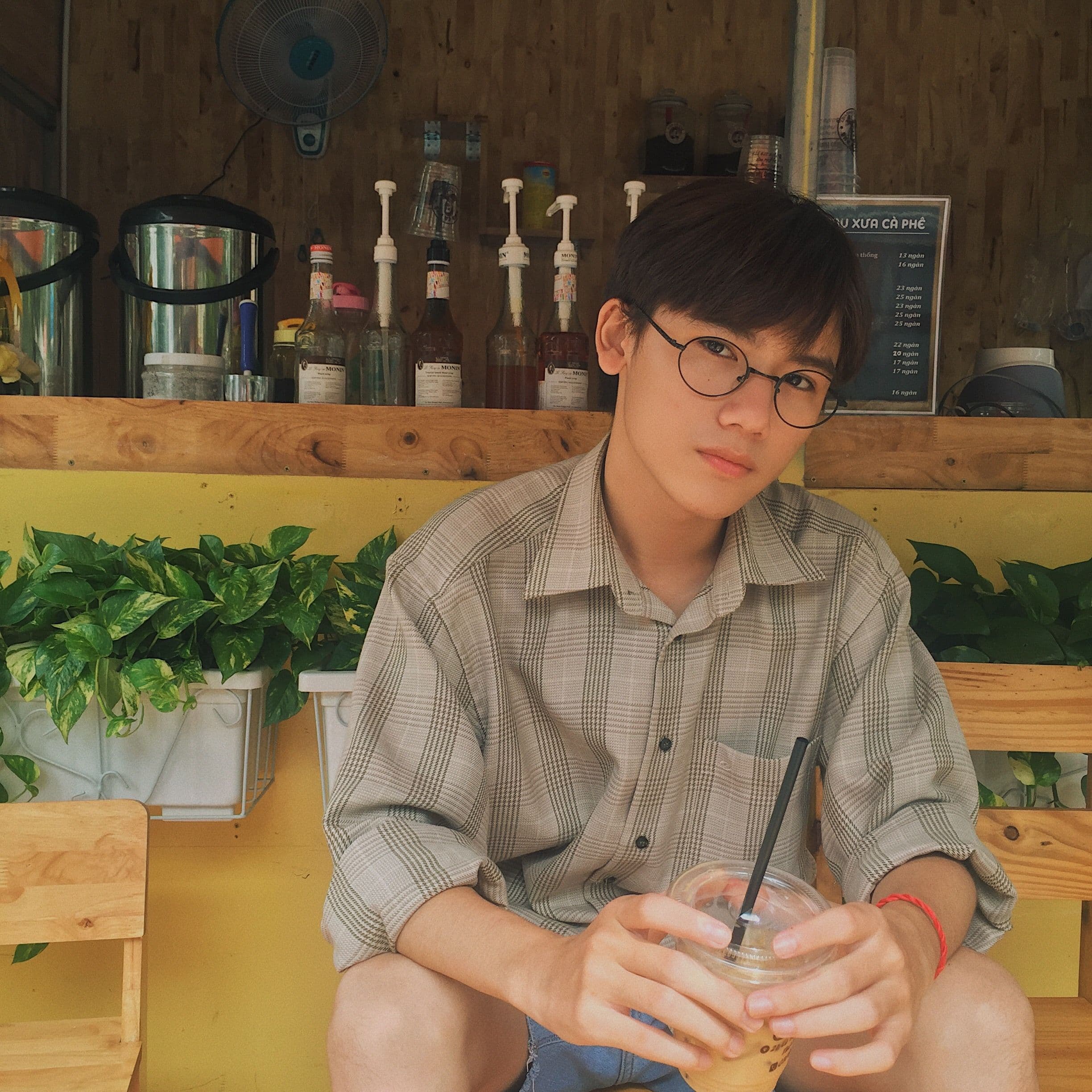 Owen’s reference photo, wearing round glasses and a checked shirt while holding a drink in a café